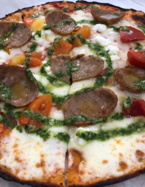 A pizza topped with sausage slices, tomatoes, cheese, and a swirl of pesto sauce on a crispy crust.