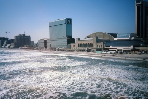A view of a beach with waves, featuring modern buildings and a boardwalk in the background under a clear blue sky.