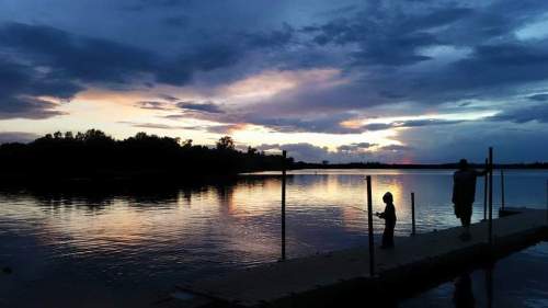 Silhouette of a child and an adult fishing on a dock at sunset, with colorful clouds reflecting on the water.
