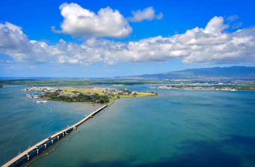 Aerial view of a bridge extending over water, with lush land and clouds in the background.