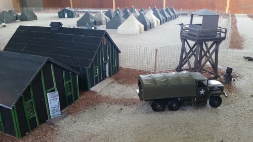 A miniature military camp scene featuring tents, a watchtower, and a truck on a gravel surface.