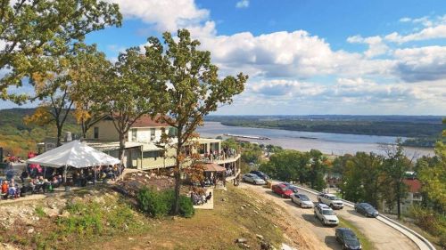A scenic view of a river from a hilltop, featuring a gathering area with people, trees, and cars along the road.