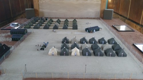 Aerial view of a miniature military camp with tents, buildings, and small figures on a gravel surface.
