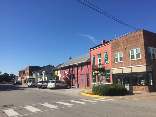 A sunny street view of a small town with colorful buildings, parked cars, and clear blue skies.