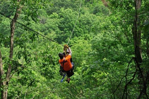 A person in an orange shirt zip-lining through a lush green forest.