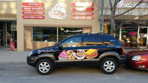 A black car parked outside a bakery, featuring colorful food graphics like cookies, cupcakes, and a sandwich.