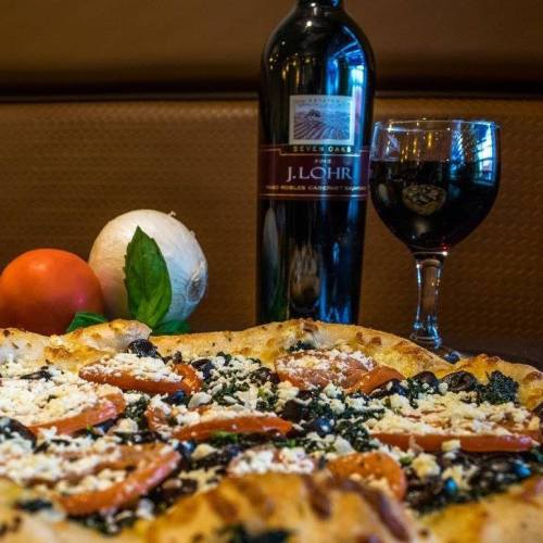 A close-up of a pizza topped with tomatoes, spinach, and cheese, accompanied by a bottle of red wine and a glass.