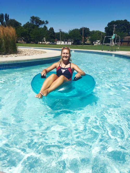 A person relaxes on a blue inner tube in a sunny pool, surrounded by grass and trees.