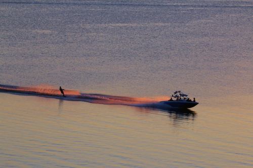 A person water skiing behind a boat on a calm lake at sunset, creating a spray of water.