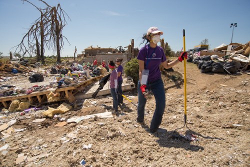 Volunteers clean up debris in a disaster-affected area, wearing masks and gloves, with piles of trash and damaged structures around.