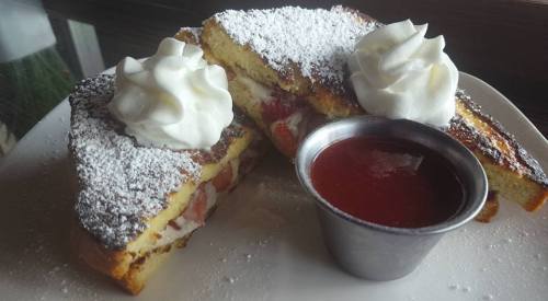 Two stuffed French toast slices topped with whipped cream and powdered sugar, served with a side of strawberry sauce.