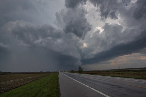 A wide road stretches through a rural landscape under a dramatic sky filled with dark, swirling storm clouds.