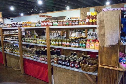 A rustic display of various jars and bottles of sauces, jams, and pickles on wooden shelves in a market setting.