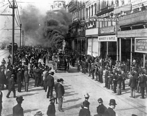 Historic street scene with crowds, smoke, and buildings, capturing a moment from the past.