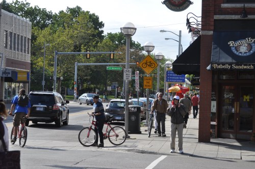 A busy street scene with cyclists, pedestrians, and shops, featuring traffic lights and a bike lane sign.