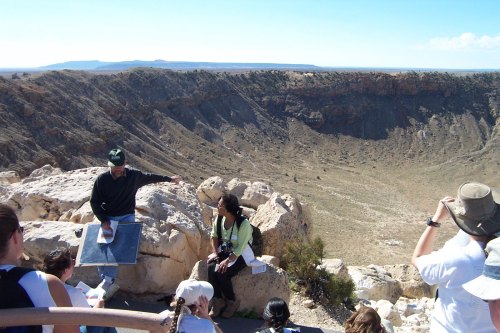 A group of people listens to a guide at a scenic overlook of a large crater in a desert landscape.