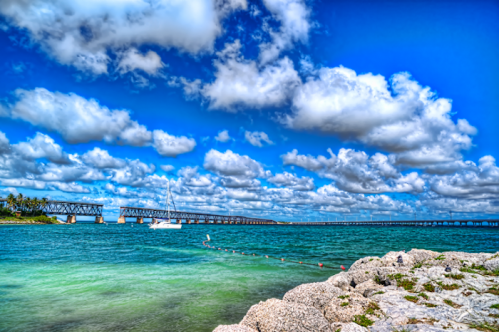 A scenic view of a blue ocean with a bridge in the background, fluffy clouds, and a sailboat near the shore.
