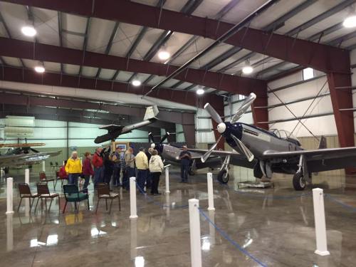 A group of people gathers around vintage aircraft in a spacious hangar with metal beams and polished floors.