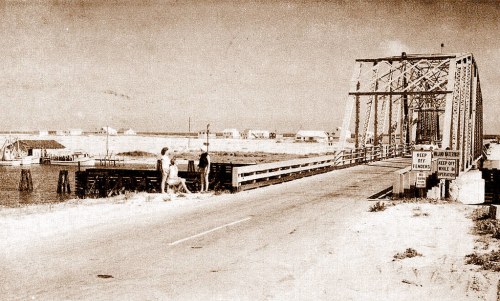 A vintage photo of a bridge with two people walking, surrounded by a rural landscape and distant buildings.