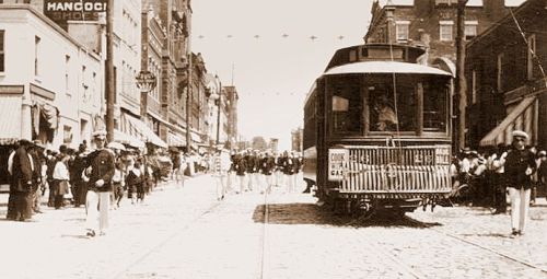 Historic street scene featuring a trolley, pedestrians, and storefronts, set in a bustling urban environment.