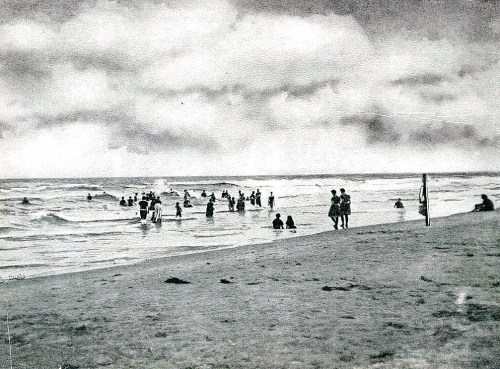 Black and white image of people swimming and playing at the beach, with waves and cloudy skies in the background.