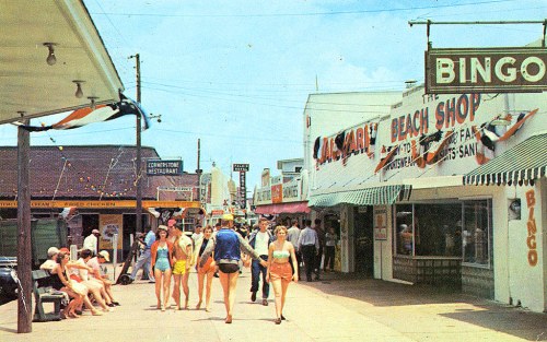 A lively beachside street scene with shops, people in swimwear, and colorful signage under a bright blue sky.