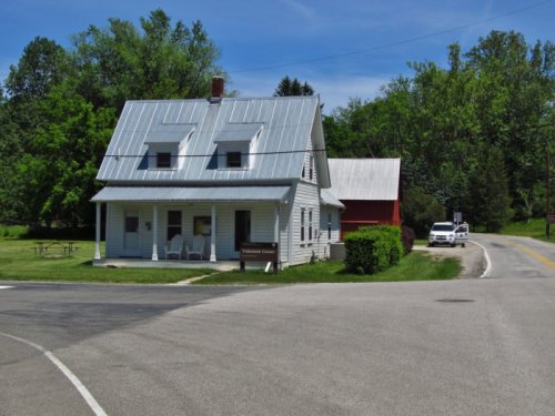 A quaint two-story house with a metal roof, surrounded by greenery and a road, featuring a sign for "Visitor Center."