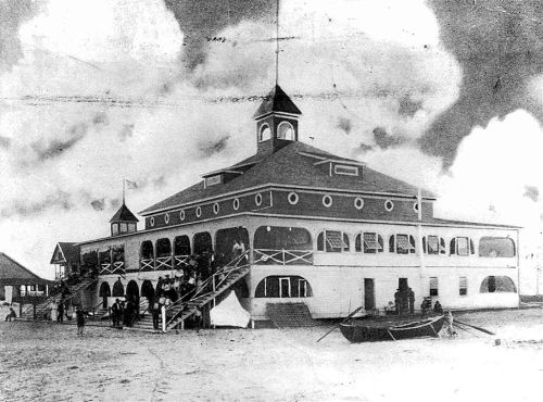 Historic black-and-white photo of a large, multi-story building with a tower, surrounded by people and boats.