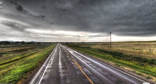 A long, empty road stretches into the distance under a cloudy, overcast sky, with wet pavement reflecting the light.