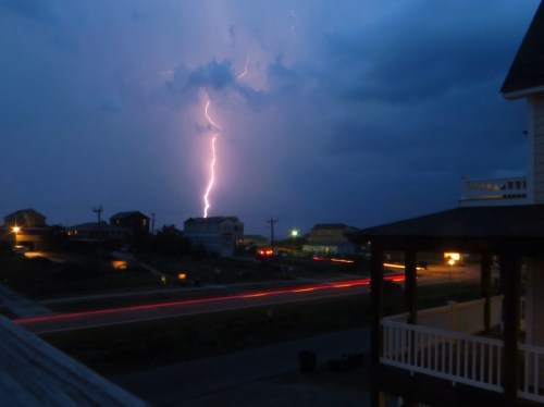 A dramatic night scene with lightning striking near houses, illuminated by streetlights and car trails on a road.