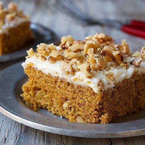 A slice of moist pumpkin cake topped with cream cheese frosting and chopped walnuts on a silver plate.