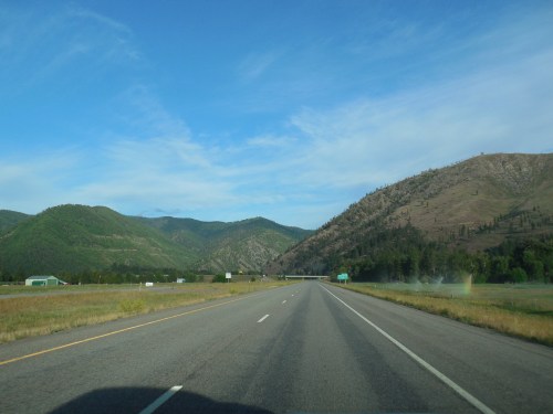A wide highway stretches through green mountains under a clear blue sky, with a few clouds in the distance.