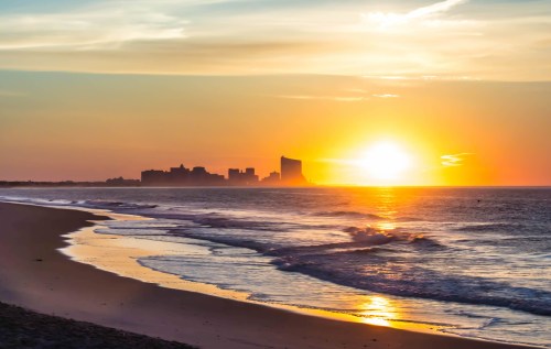 Sunset over a beach with gentle waves, reflecting golden hues, and a city skyline silhouetted against the horizon.