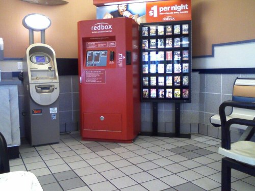 A Redbox movie rental kiosk and an ATM in a small, tiled waiting area with chairs.