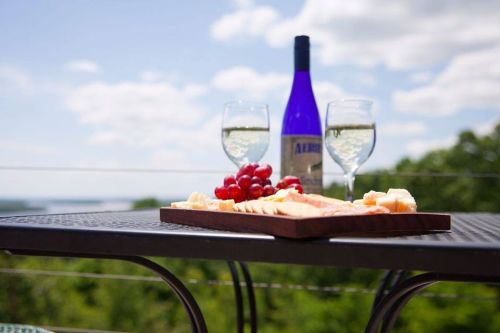 A table set with a cheese platter, grapes, and two glasses of white wine, with a bottle in the background and a scenic view.