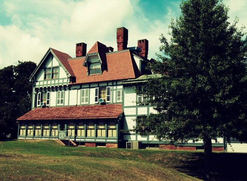 A large, historic house with a steep roof, multiple chimneys, and a grassy area in front, under a partly cloudy sky.
