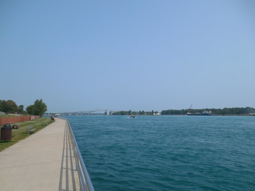 A serene waterfront view with a walkway, trees, and a distant bridge under a clear blue sky.