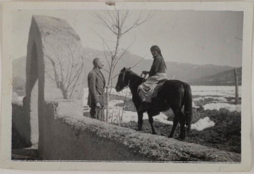 A woman on a horse talks to a man near a stone arch, with mountains and snow in the background.