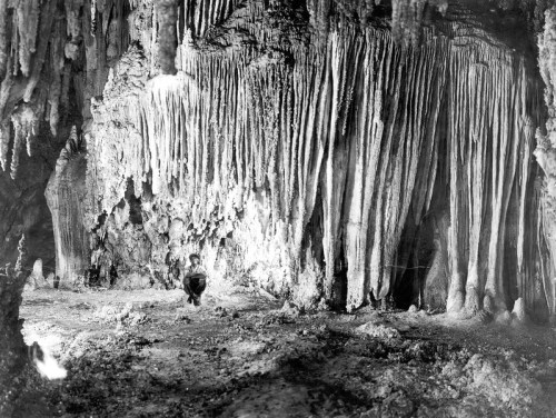 A black and white photo of a cave with stalactites and a person sitting on the ground, surrounded by rocky formations.