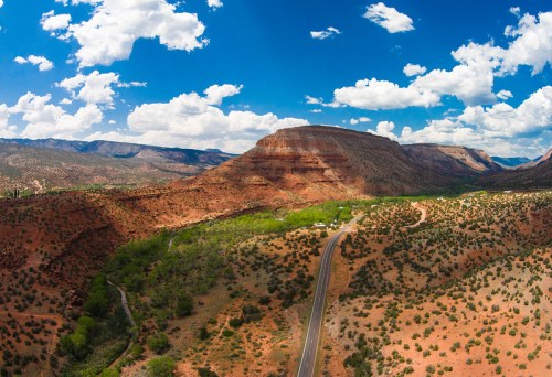 A scenic view of red rock formations and a winding road under a bright blue sky with fluffy white clouds.