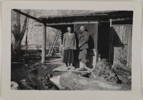 A black-and-white photo of a man and woman standing outside a rustic cabin, surrounded by trees and logs.