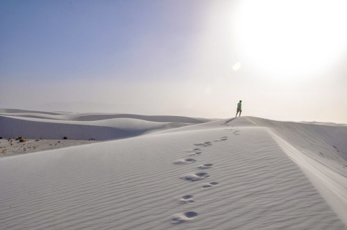 A person stands on a sandy dune under a bright sun, with footprints leading across the smooth, white landscape.