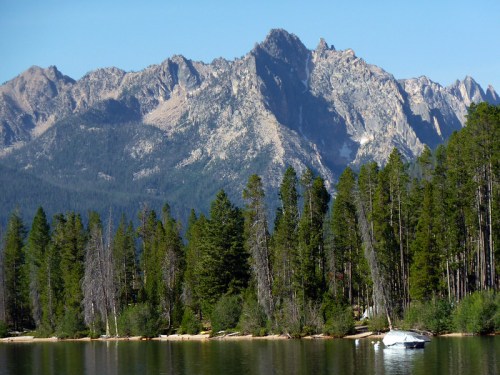 A serene lake surrounded by tall trees, with majestic mountains rising in the background under a clear blue sky.
