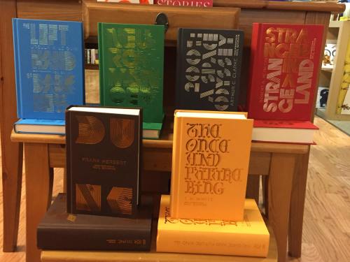 A collection of colorful book covers displayed on a wooden table, featuring titles like "Dune" and "2001: A Space Odyssey."