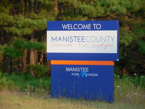 Welcome sign for Manistee County, Michigan, surrounded by trees, promoting the area's natural beauty.
