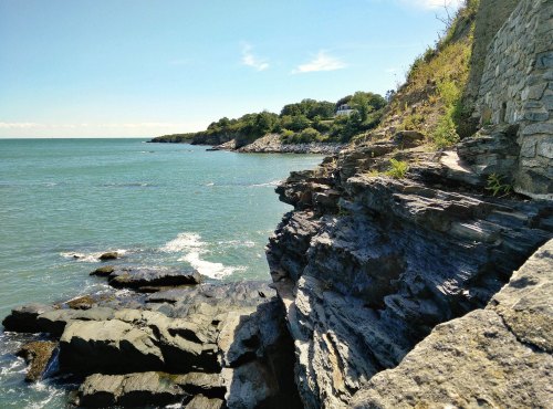 Rocky coastline with clear blue water and greenery in the background under a bright, sunny sky.