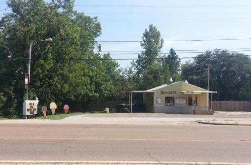 A small ice cream shop with a yellow exterior, surrounded by trees, and a gravel parking area on a sunny day.