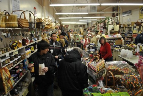 A busy store interior filled with shoppers browsing various goods, including baskets, candy, and jars of preserves.