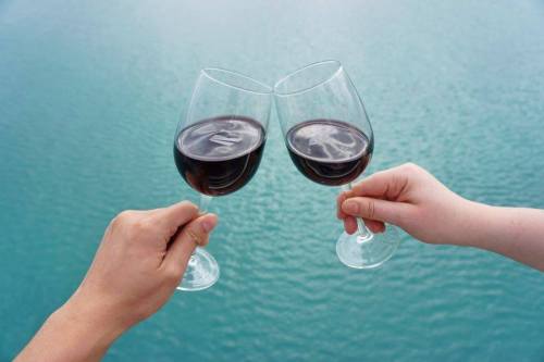 Two hands holding wine glasses, clinking them together against a backdrop of calm blue water.