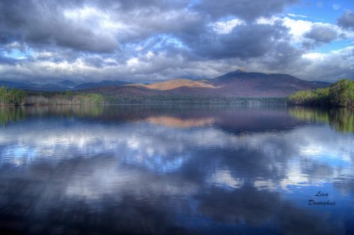 A serene lake reflects mountains and clouds, surrounded by trees in vibrant autumn colors.
